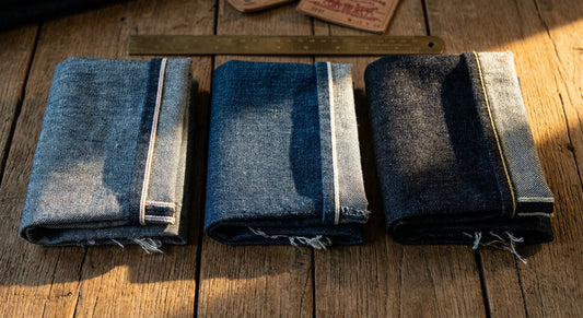 Three folded selvedge denim fabric swatches of varying weights arranged side by side on a wooden surface, lit dramatically to reveal weave texture and selvedge edge detail.