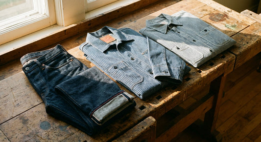A flat lay of raw indigo selvedge denim jeans alongside a Wabash stripe chore coat and hickory stripe shirt arranged on a weathered wooden surface in warm natural light.