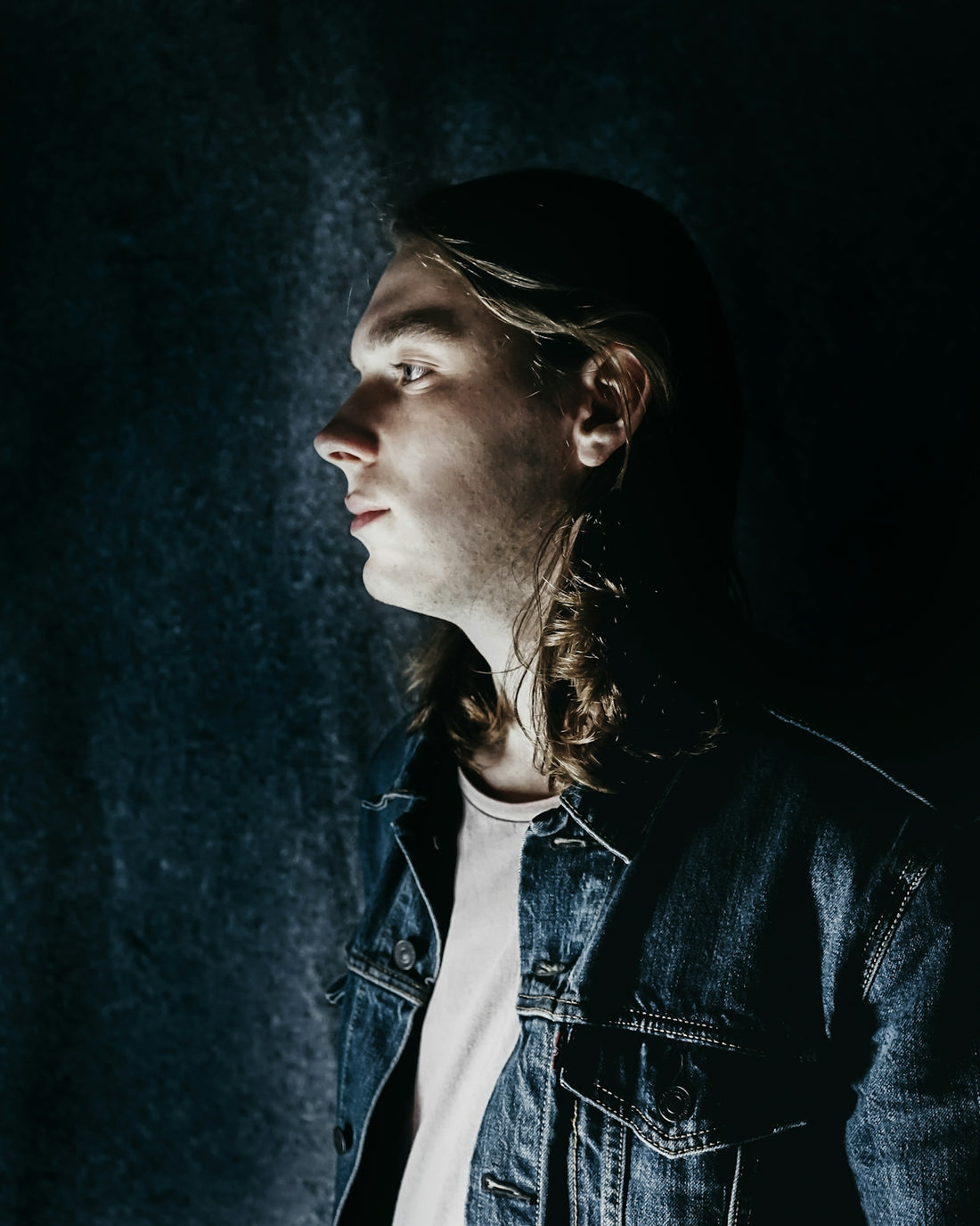 man wearing blue denim jacket standing in front of black wall