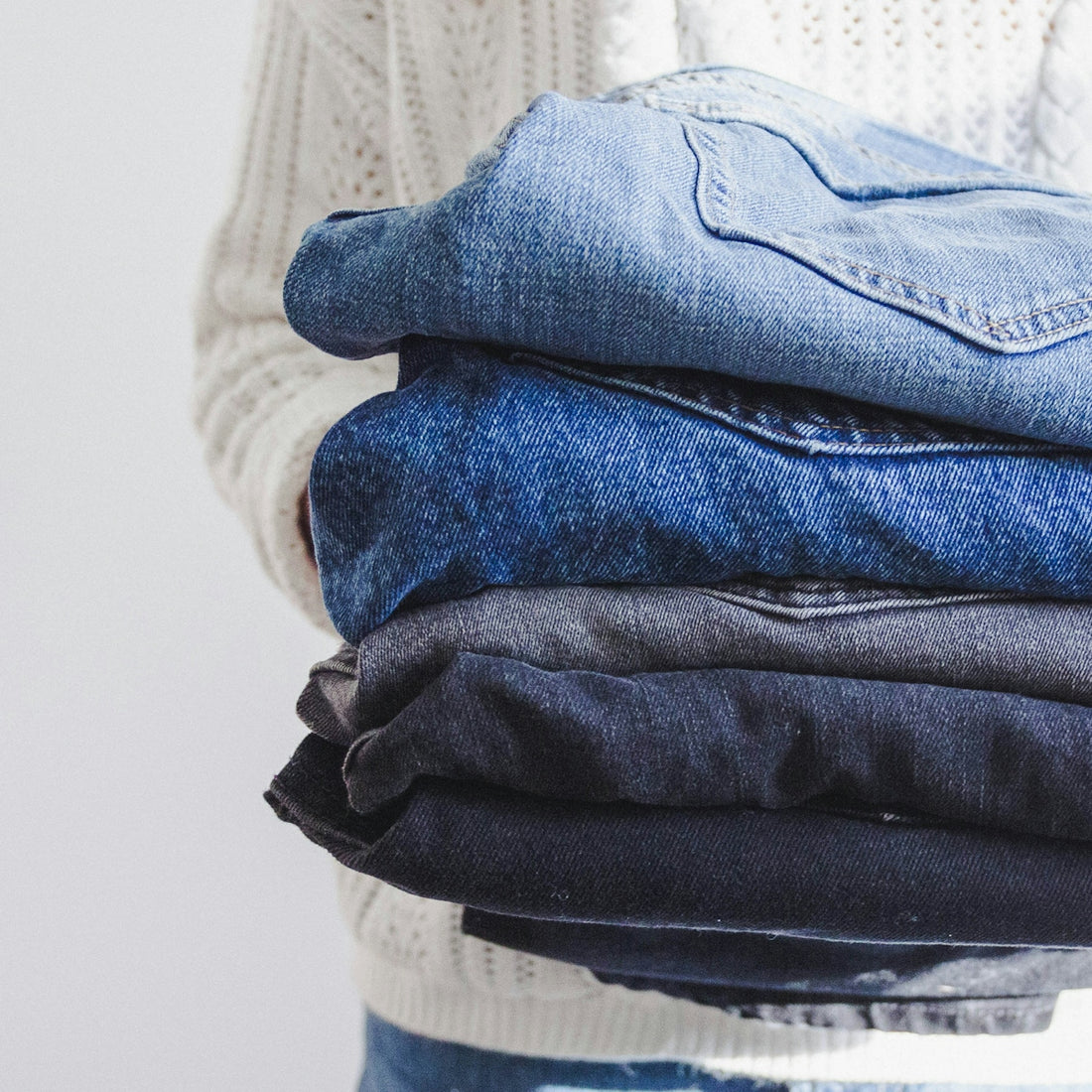 A person in a white cable-knit sweater holding a stack of folded denim jeans