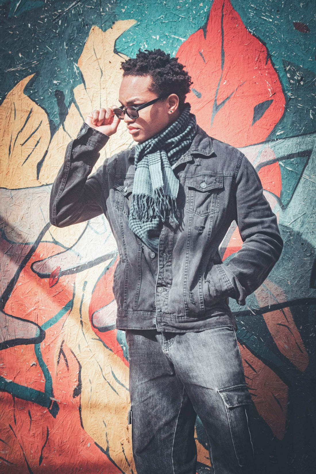 Man in denim jacket and scarf poses by graffiti wall