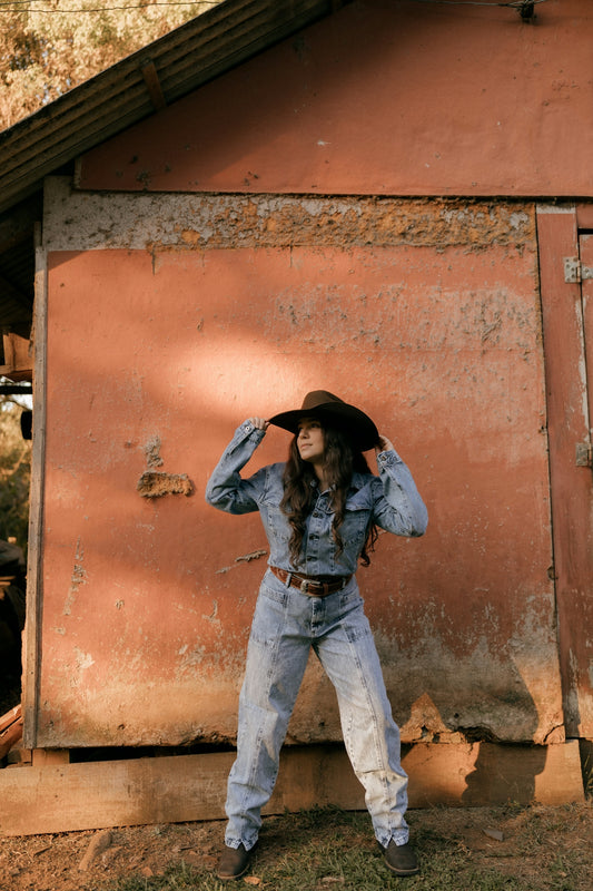 Woman in denim outfit and cowboy hat poses near barn