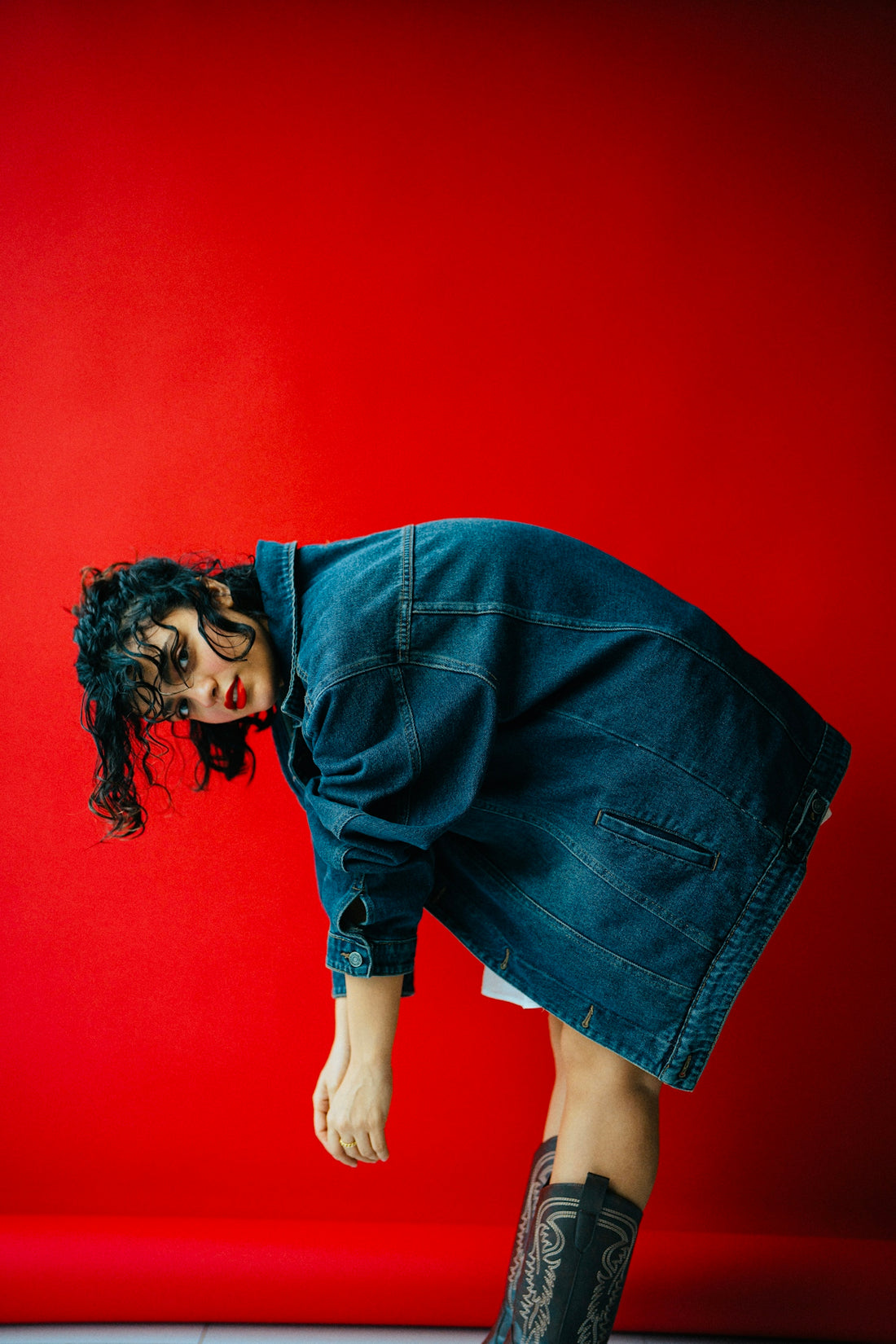 Woman in denim jacket bends over red backdrop